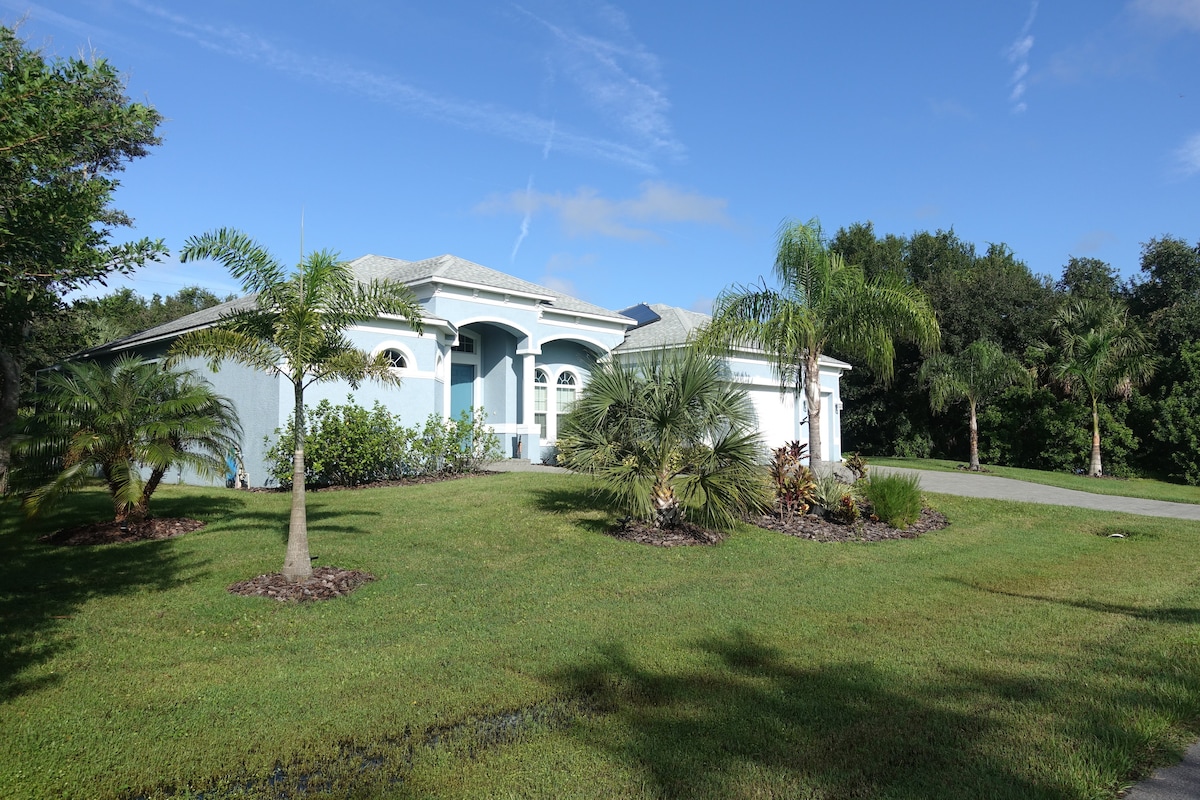 The exterior of a contemporary home is showcased, featuring a light blue facade with arched windows and a spacious entryway. Lush green landscaping and palm trees enhance the tropical atmosphere, while a well-maintained lawn surrounds the property, reflecting a serene outdoor setting.