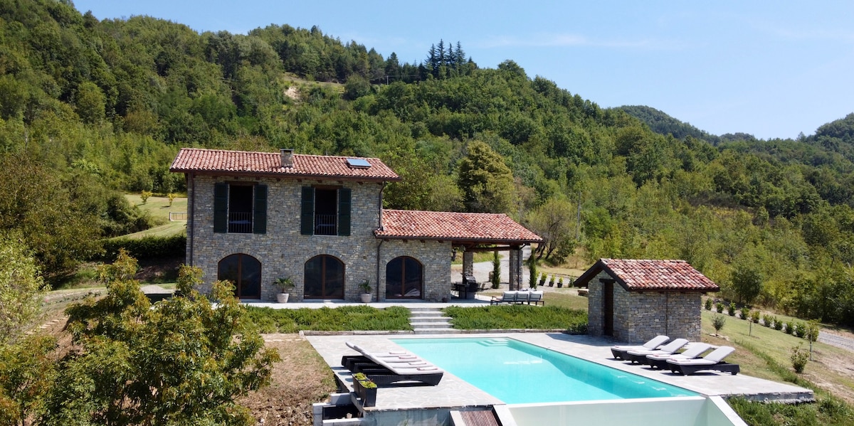 A spacious two-story stone house is surrounded by lush greenery and rolling hills. A well-maintained outdoor area features a large pool with sun loungers, complemented by a nearby pool house. The terracotta roof adds a rustic touch to this inviting setting.