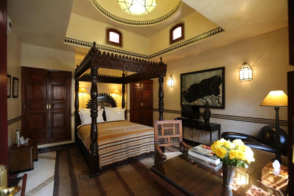 A room features a traditional canopy bed with decorative woodwork, complemented by warm-colored walls and a geometric ceiling light. A wooden chair and a small table are positioned near a vase of flowers. A TV and cabinetry are situated against one wall, enhancing the overall design.