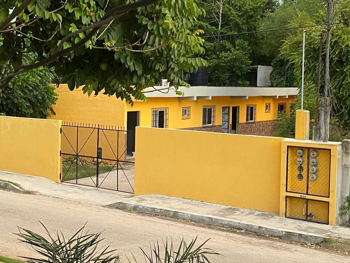 A vibrant yellow building is enclosed by a gated wall, featuring a black wrought-iron gate. The property showcases several windows, and the entrance is visible beyond the gate, surrounded by greenery and a neatly landscaped front yard.