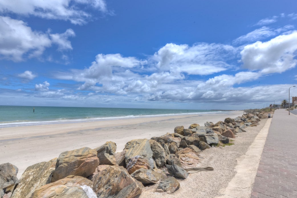A wide view of the beachfront extends under a clear blue sky, with fluffy clouds enhancing the scenic horizon. The sandy shoreline is lined with smooth rocks, providing a natural border to the beach. The tranquil sea gently laps at the shore.