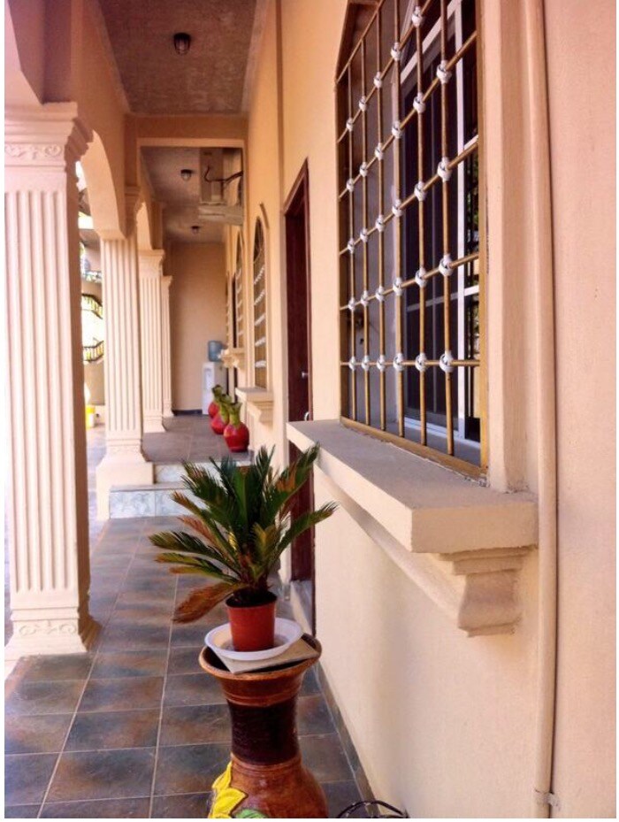 A shaded walkway features decorative columns and tiled flooring. Potted plants add greenery along the path, while windows with iron grilles provide natural light. The entrance to a room is visible, framed by a door that complements the warm tones of the exterior.