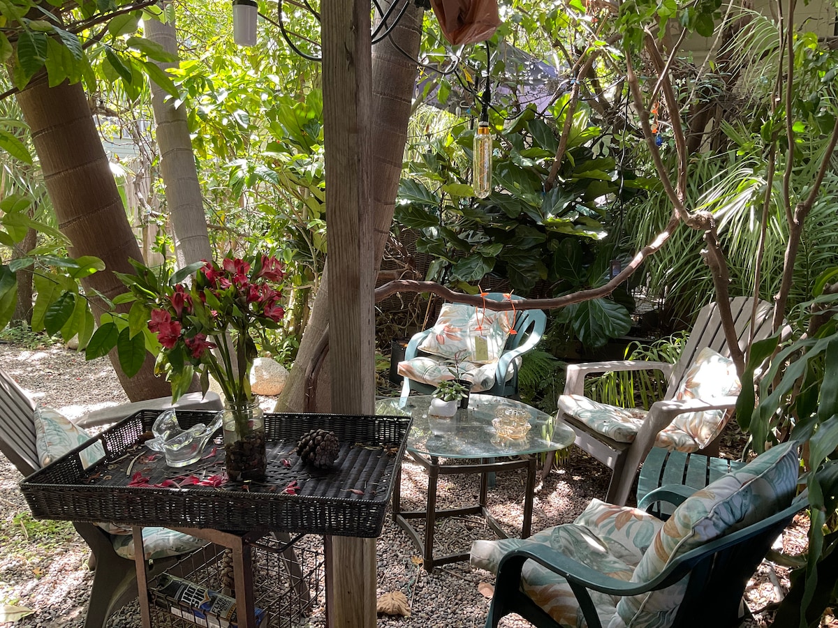 An outdoor space features a blend of seating options, including a glass-topped table and cushioned chairs. Surrounding tropical plants and trees create a lush, private atmosphere. A floral centerpiece and decorative items enhance the serene environment, inviting relaxation and connection with nature.