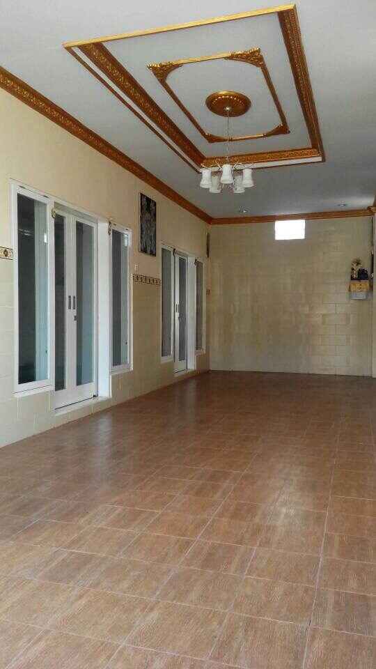 A spacious room features a light-colored tiled floor and elegant molding along the ceiling. Large windows allow natural light to enter, with a small window positioned higher on the wall. A simple chandelier provides additional lighting in the center of the room.
