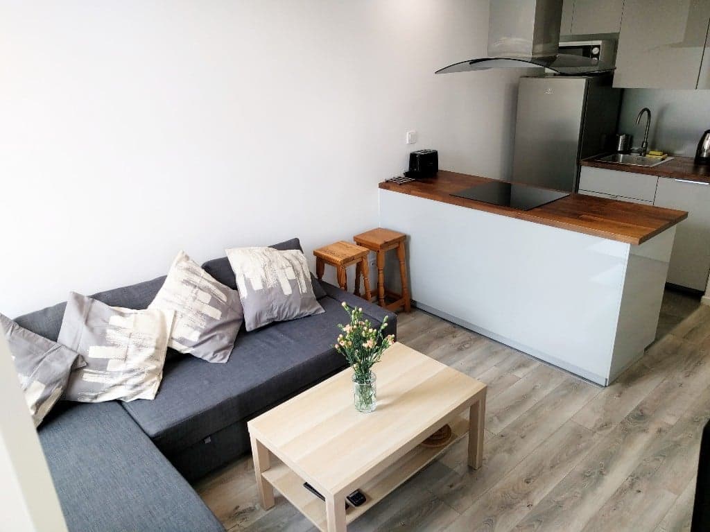 A modern living area is presented with a gray sectional sofa adorned with decorative pillows. A wooden coffee table sits in front, complemented by a small vase with fresh flowers. The kitchen area, featuring a countertop with bar stools and stainless steel appliances, is visible in the background.