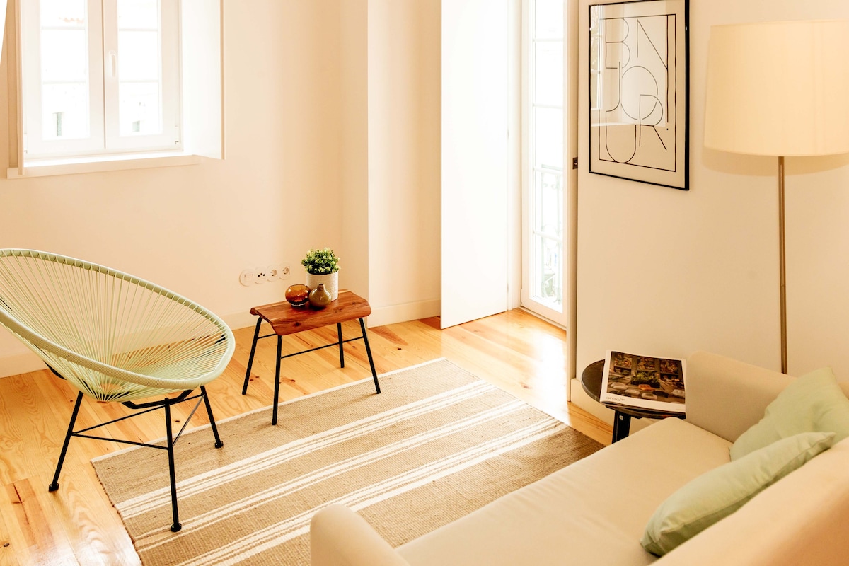 A light-filled living area features a comfortable sofa and an acapulco chair. A small wooden table is positioned between them, complemented by a potted plant. The warm wooden floors enhance the bright ambiance, while a framed art piece catches the eye on the wall.