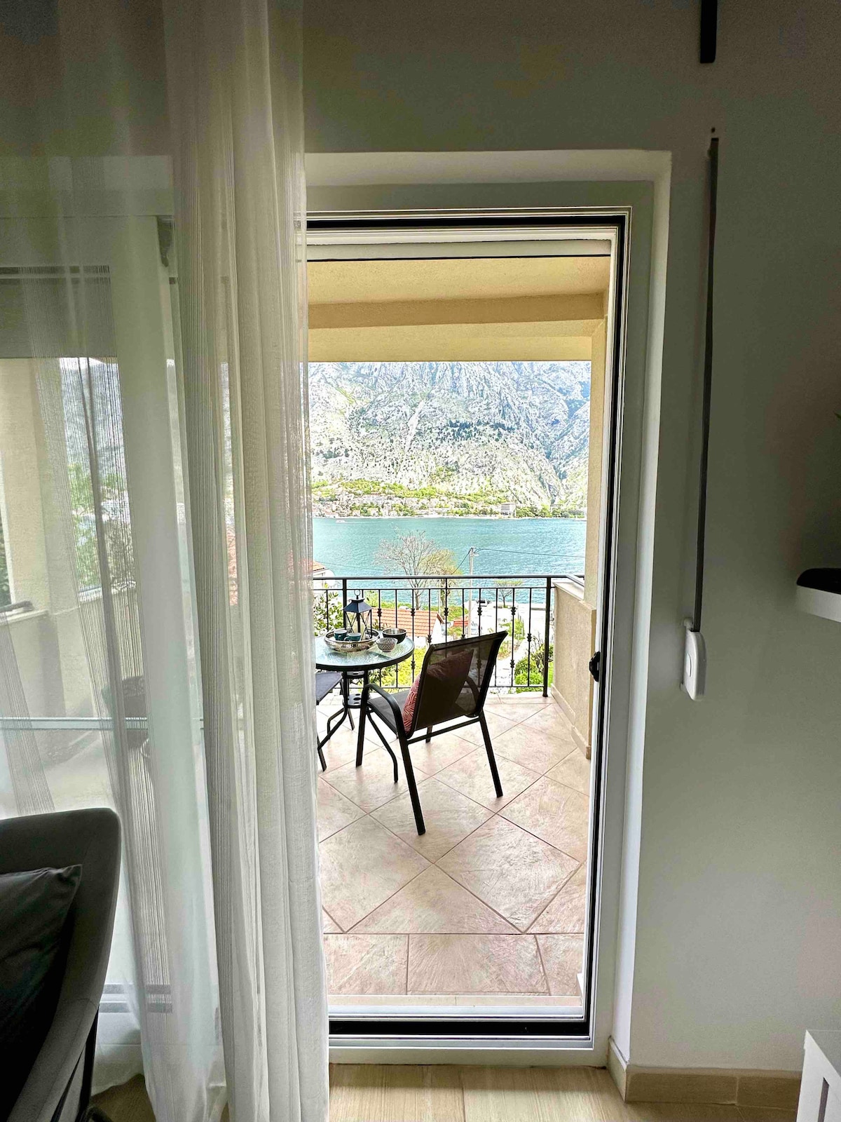 A door opens to a terrace with a view of the tranquil waters of Boka Bay. A small table and two chairs are positioned on the balcony, framed by a glass door and sheer curtains, inviting relaxation while enjoying the surrounding natural beauty.