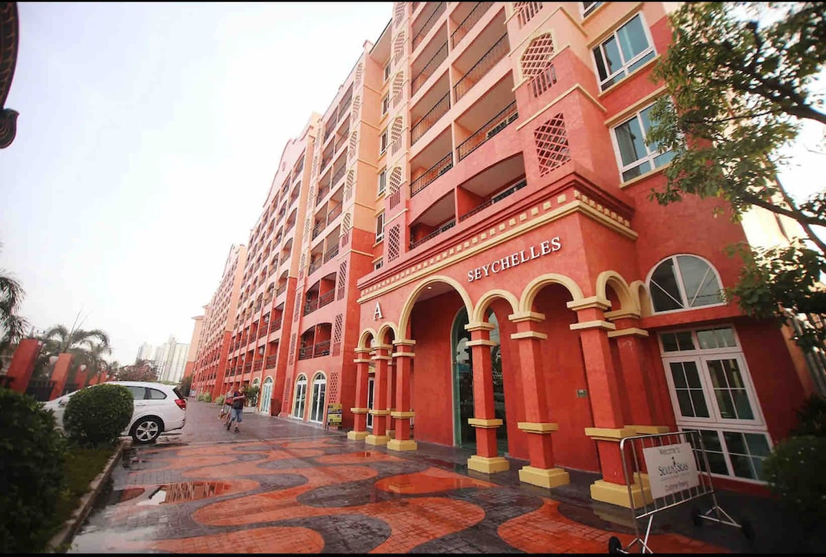 A colorful condominium complex is viewed from the exterior, showcasing a symmetrical facade painted in shades of red and cream. The entrance features arched details with the name 'Seychelles' elegantly displayed above. Pathways lined with greenery lead toward the building in the foreground.