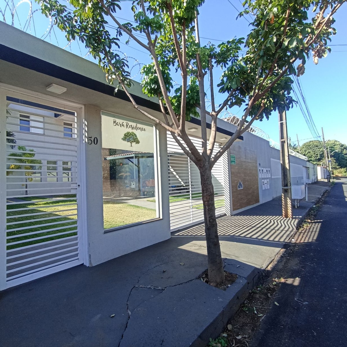 The exterior of the property features a modern facade, highlighted by a large window displaying the inviting interior. A small tree provides shade in front, while the paved walkway leads to the entrance. Clear blue skies are visible overhead, completing the serene setting.