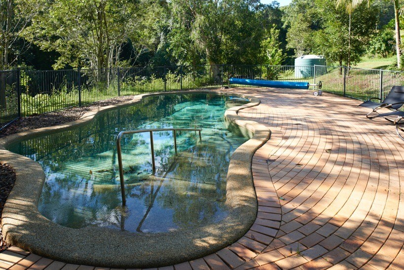 An in-ground pool features a gently curving shape, surrounded by a spacious sandstone patio. Lush greenery provides a natural backdrop, while a safety fence encloses the area. A pool ladder is positioned for easy access. A water tank is visible in the distance.