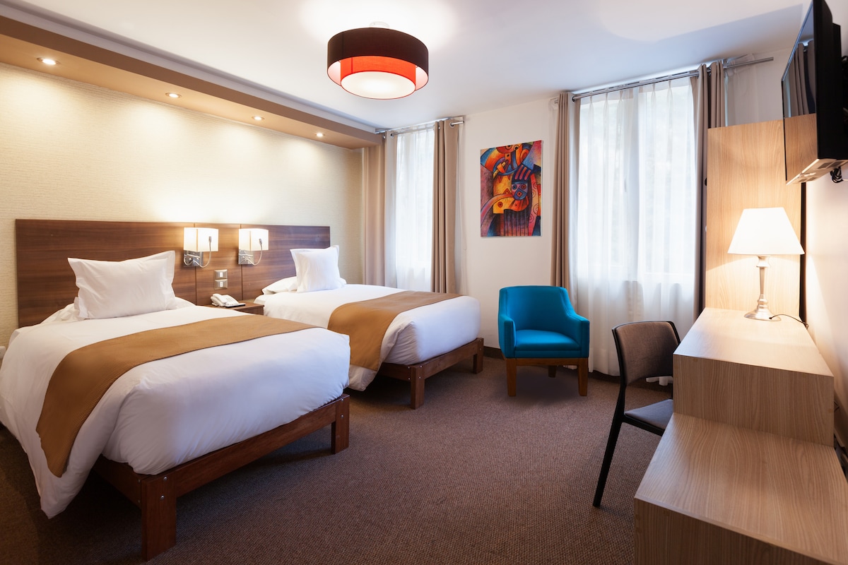 The room features two spacious beds dressed with white linens and brown accents. Natural light filters through sheer curtains, illuminating the decor, which includes a colorful wall art piece. A small blue chair and a simple desk with a lamp complete the inviting space.