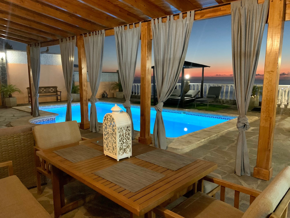 A spacious outdoor terrace is framed by wooden beams and draped curtains, providing a relaxing seating area. A wooden table sits under soft lighting, while a turquoise pool reflects the evening sky. Comfortable loungers are positioned nearby, inviting relaxation.