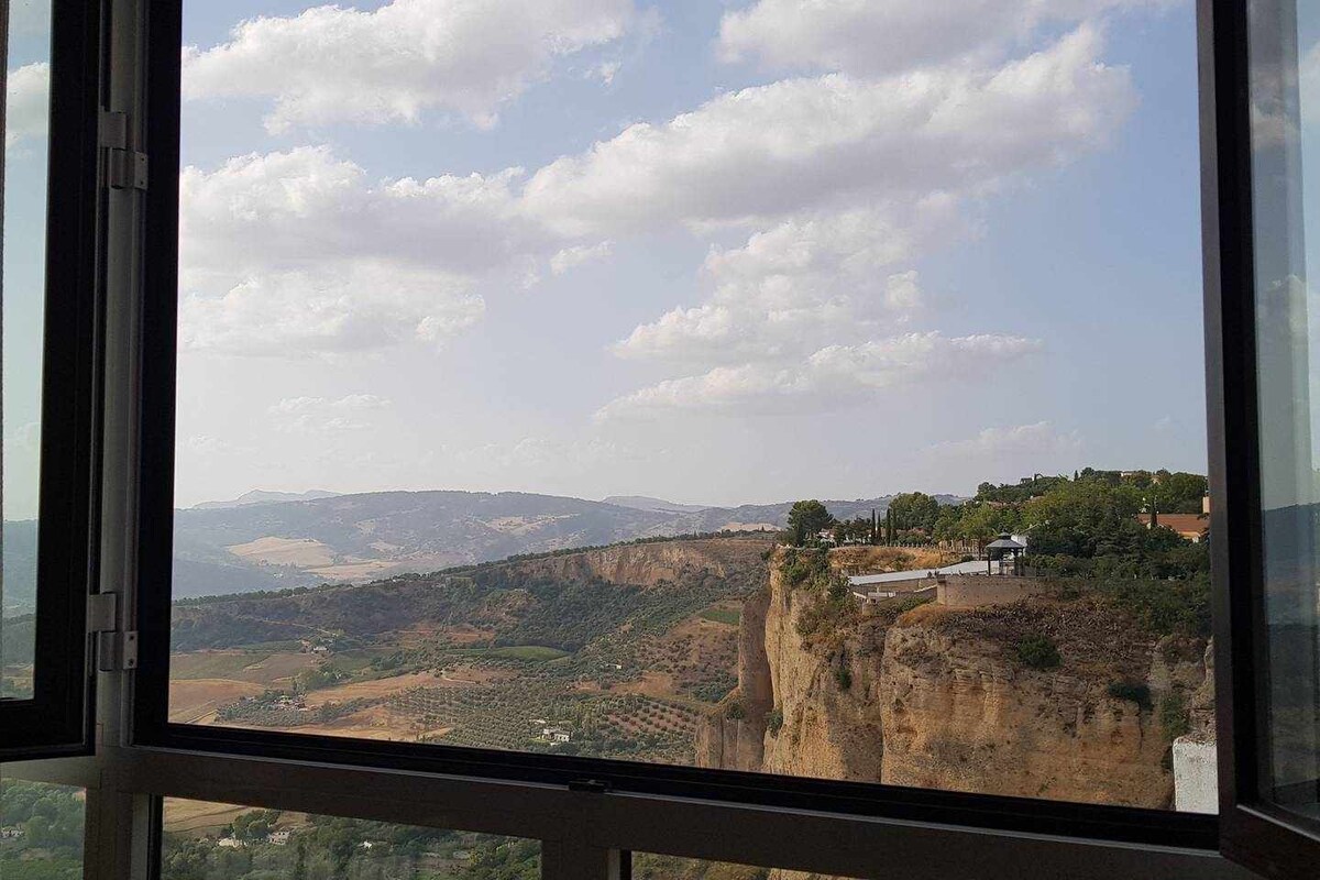 A view of rolling hills and valleys extends from the large window, framed by clear glass. Soft clouds drift across the sky, while lush greenery can be seen in the distance, capturing the natural beauty of the surrounding landscape.