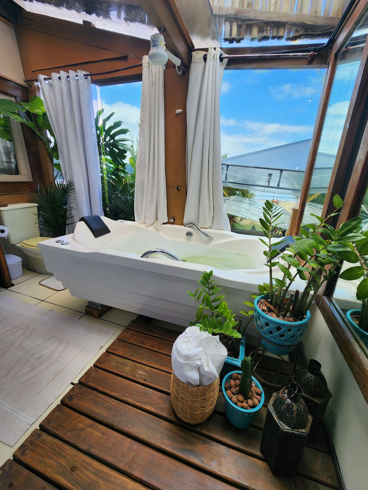A modern bathroom is illustrated with a freestanding bathtub positioned beside floor-to-ceiling windows, allowing natural light to fill the space. Potted plants add a touch of greenery, while wooden flooring contrasts with the white elements. Soft towels are neatly arranged nearby.