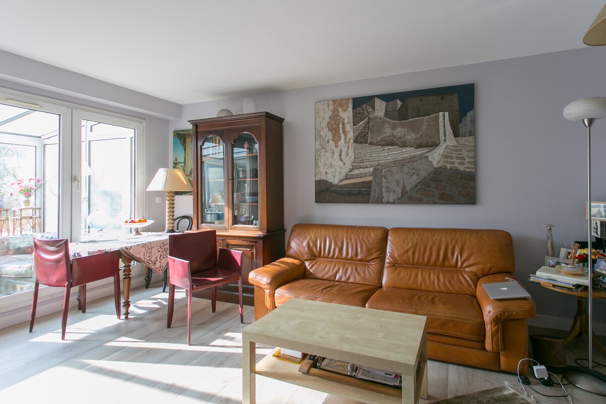 The living area is furnished with a brown leather couch and a wooden coffee table. A glass table with chairs is visible near a window, providing natural light. An artwork is displayed on the wall, and a cabinet occupies the corner, adding practical storage.