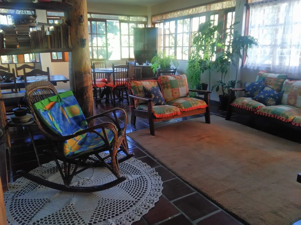 The living area features a combination of comfortable seating, including a rocking chair and sofas with bright cushions. Large windows allow natural light to flood the space, and decorative plants add a sense of tranquility. A crocheted rug lies on the tiled floor, complementing the warm atmosphere.