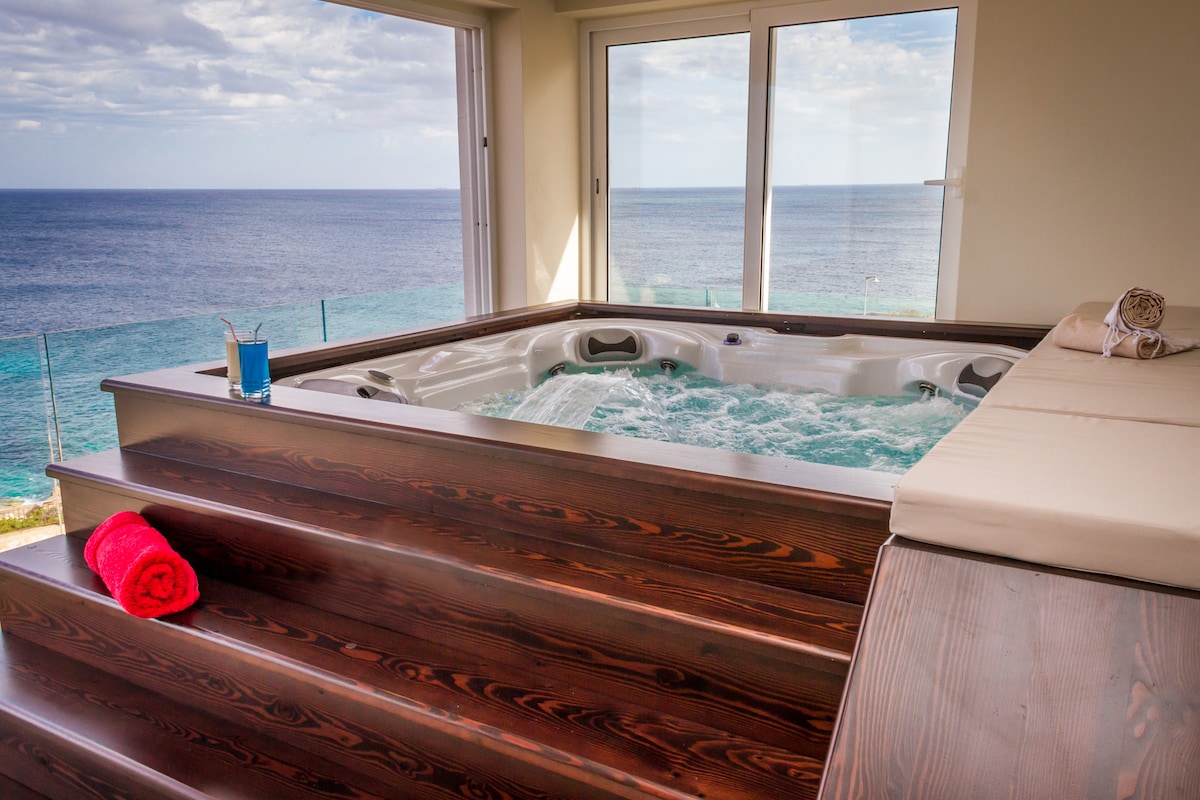 A luxurious hot tub Jacuzzi is positioned by large windows, offering unobstructed sea views. Wooden steps lead up to the tub, with a rolled red towel placed on the edge. A glass of a refreshing drink rests nearby, enhancing the relaxing ambiance.