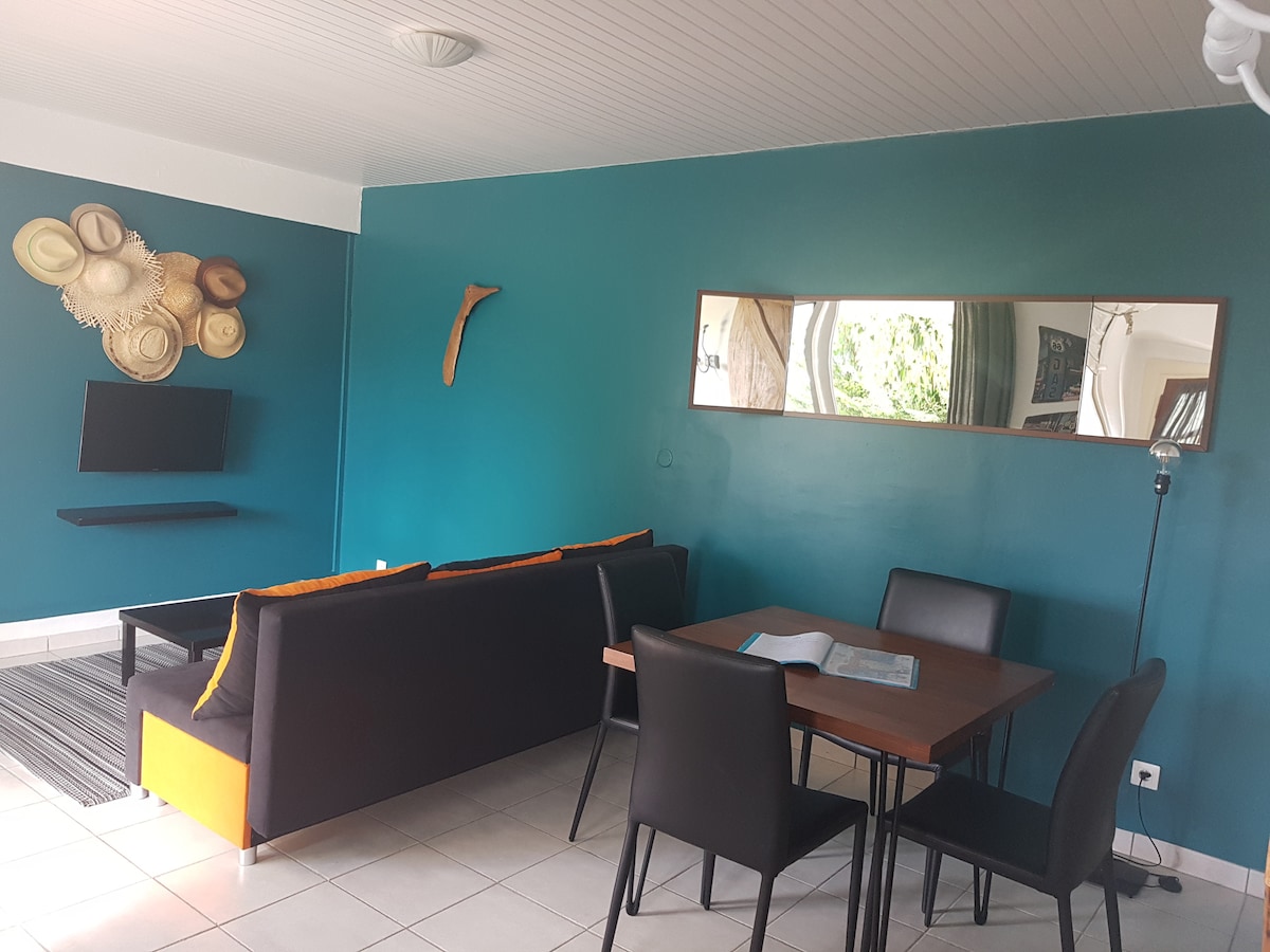 A bright living area features a couch in contrasting colors and a dining table that seats four. Wall-mounted decorations are visible, alongside a television. Large windows allow for natural light, complementing the teal walls and tiled floor.