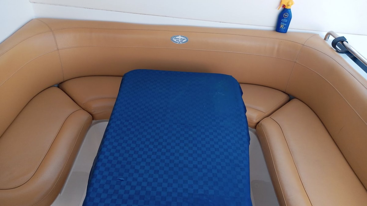 A sectional seating area is visible, featuring tan-colored upholstery. A deep blue blanket is spread over a central surface, complementing the cozy atmosphere. A sunblock bottle is positioned in the background, indicating a relaxing space for sunbathing.