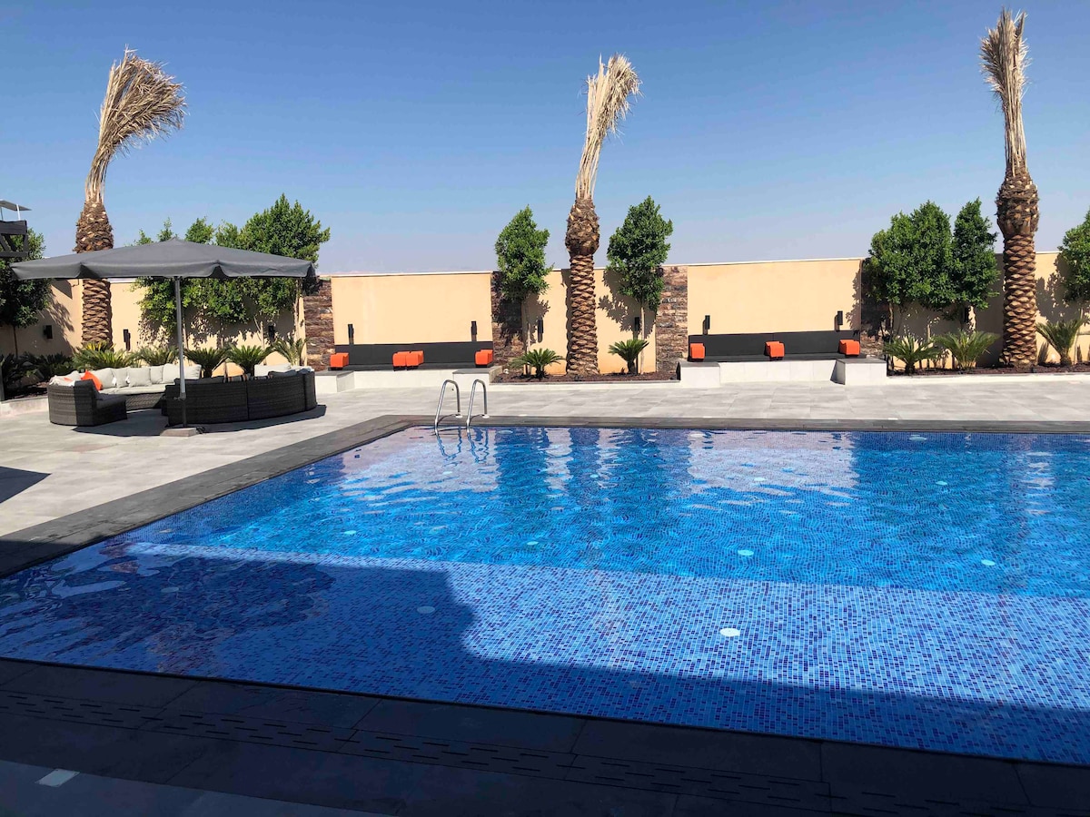 An expansive swimming pool of clear blue water is framed by palm trees and lounge seating. The pool area features shaded spaces with comfortable seating, creating an inviting outdoor environment for relaxation.