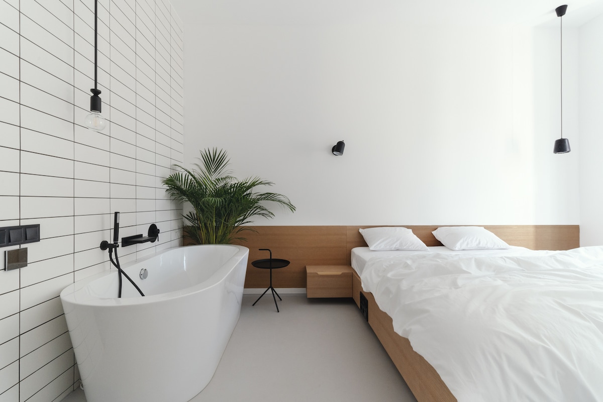 A modern bedroom features a spacious bed with white linens against a minimalist backdrop. A freestanding bathtub sits beside the bed, while subtle greenery adds a touch of nature. Pendant lights provide soft illumination, complemented by a sleek side table.