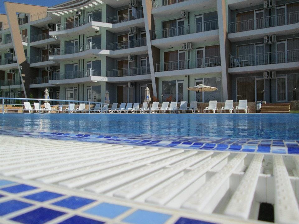 A large swimming pool is featured in the foreground, with a stylish tiled edge. Sun loungers are arranged neatly along the pool's edge, and buildings can be seen in the background, showcasing balconies and air conditioning units.