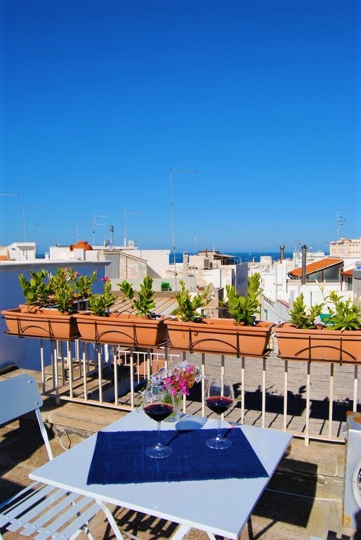 A terrace is shown, featuring a table set for two with glasses of red wine. Flower pots filled with greenery line the railing, while a clear blue sky is visible above. In the distance, the sea can be glimpsed over the rooftops of whitewashed buildings.
