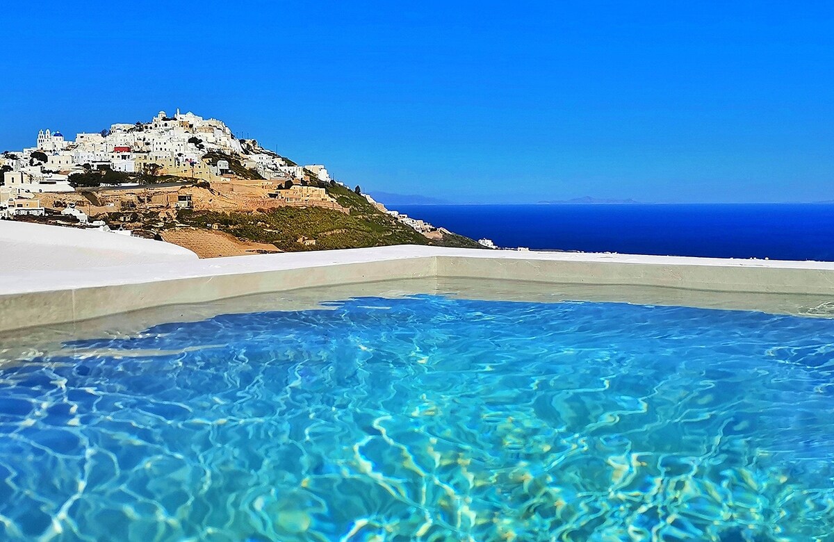 A clear blue swimming pool is framed by a smooth stone edge, reflecting the bright sky. In the background, the white buildings of a hillside village can be seen against the vibrant ocean, creating a scenic contrast with the tranquil water.