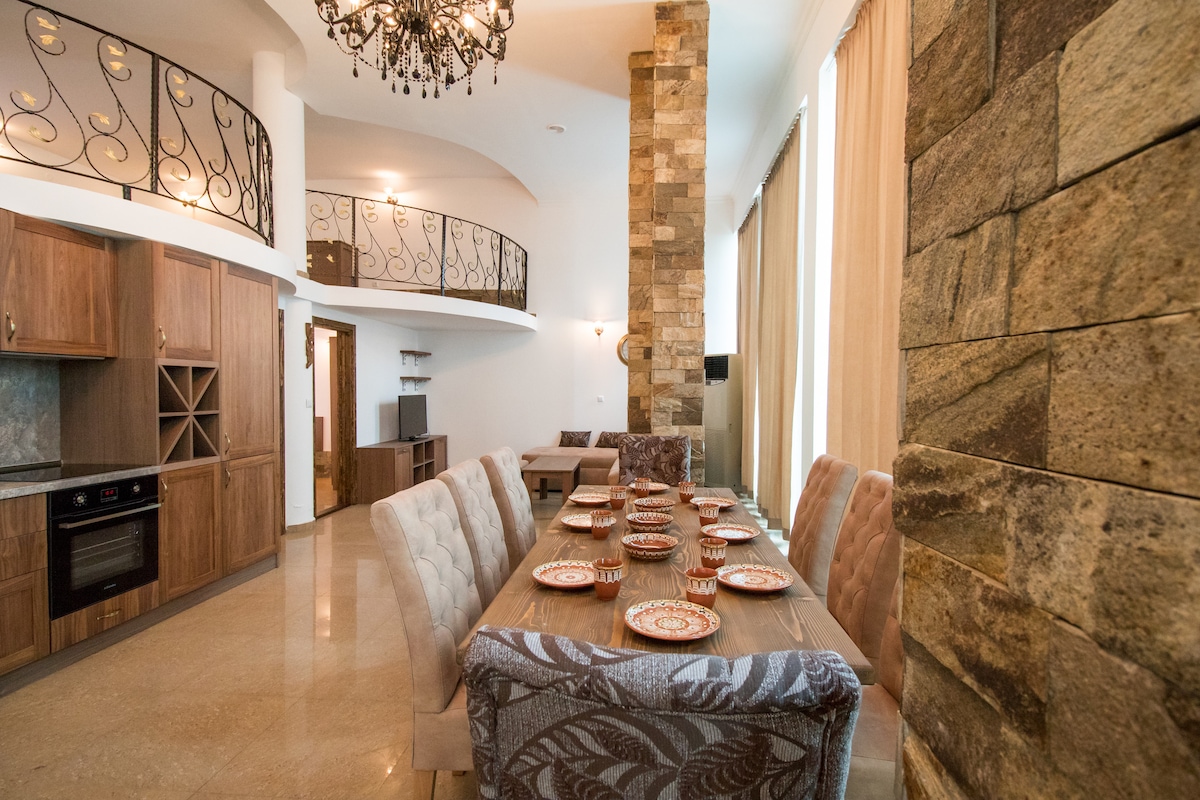 The dining area features a large table set for eight, surrounded by elegantly upholstered chairs. An open kitchen is visible to one side, equipped with modern appliances, while stone accents adorn the walls, complementing the high ceilings and large windows that allow natural light to fill the space.