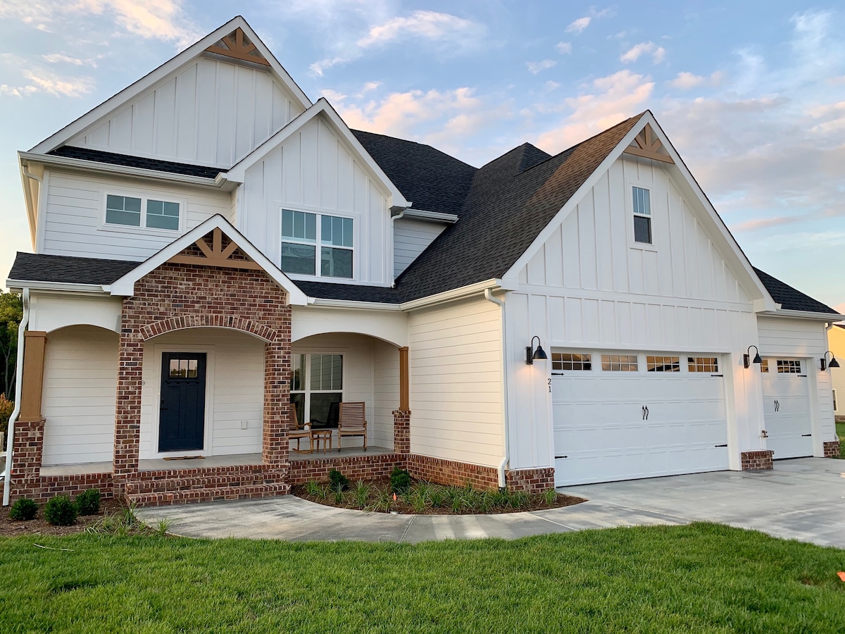 The exterior of a two-story farmhouse style home is displayed, featuring white wooden siding with brick accents. A welcoming front porch with seating creates an inviting entrance. A three-car garage is visible, complemented by a well-maintained lawn and landscaped surroundings.