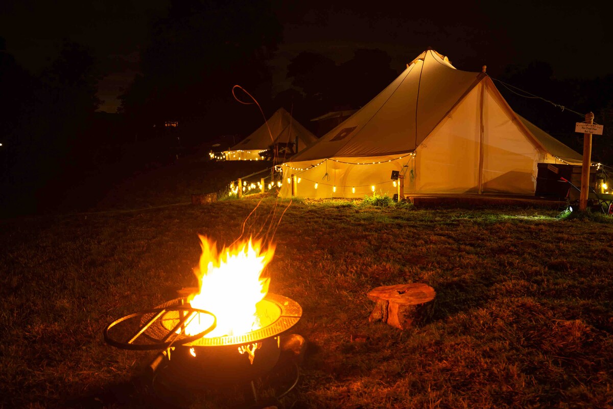 A cozy campfire burns brightly in the foreground, illuminating the area. Soft lights outline a spacious tent in the background, creating a warm ambiance. The grassy setting offers a natural camping feel, while a wooden stump sits nearby, inviting relaxation beside the fire.
