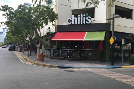 A vibrant restaurant exterior is depicted, featuring a prominent sign that reads 'chili's.' The building is characterized by an awning in red and green, with a glass railing surrounding the patio area. Nearby trees and parked cars are visible along the street.