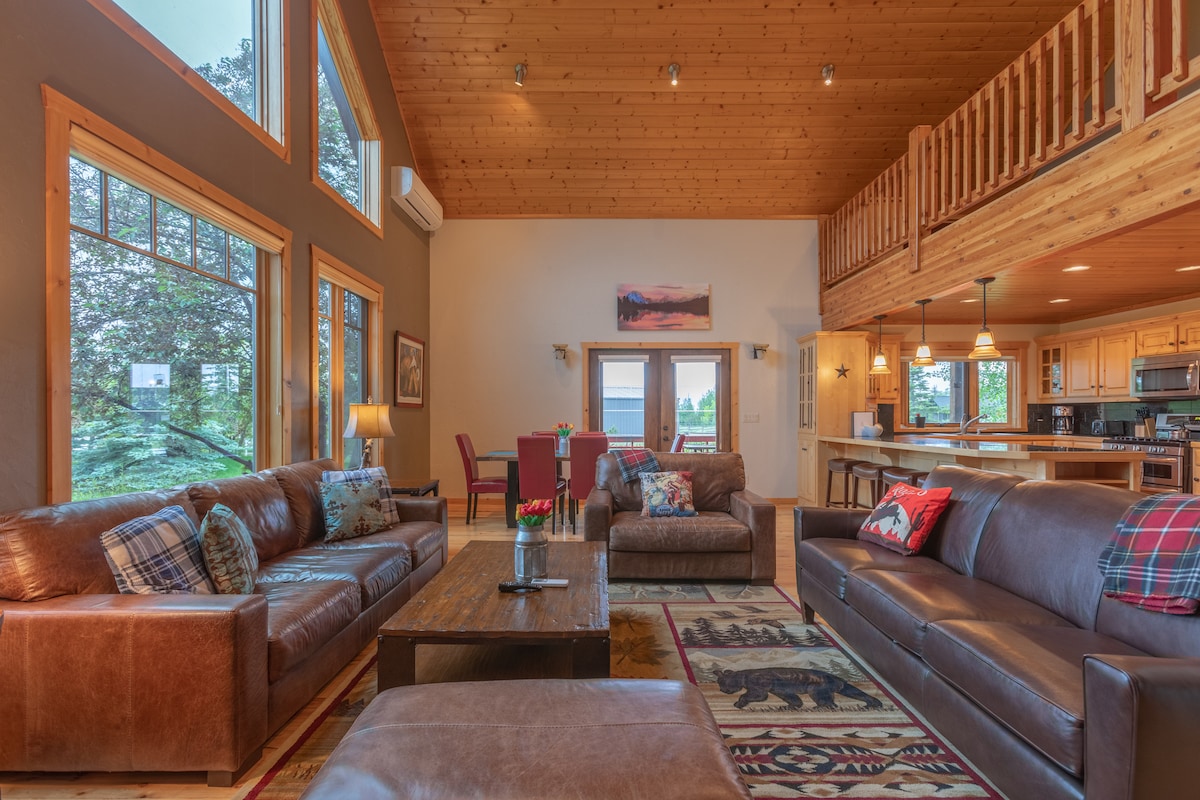 A spacious great room features high ceilings with wood paneling and large windows that allow natural light. Comfortable leather sofas are arranged around a wooden coffee table, with a decorative rug below. The open layout leads to the kitchen area, visible through an archway.