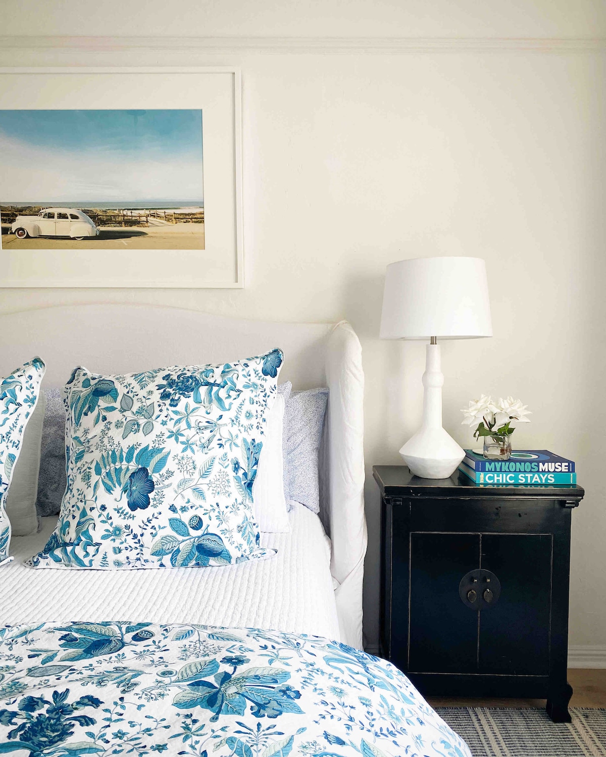 A cozy sleeping area features a light-colored headboard with a quilted design and decorative pillows in blue floral patterns. A bedside table holds a small white lamp and a vase with fresh flowers, alongside a stack of books, offering a serene touch.