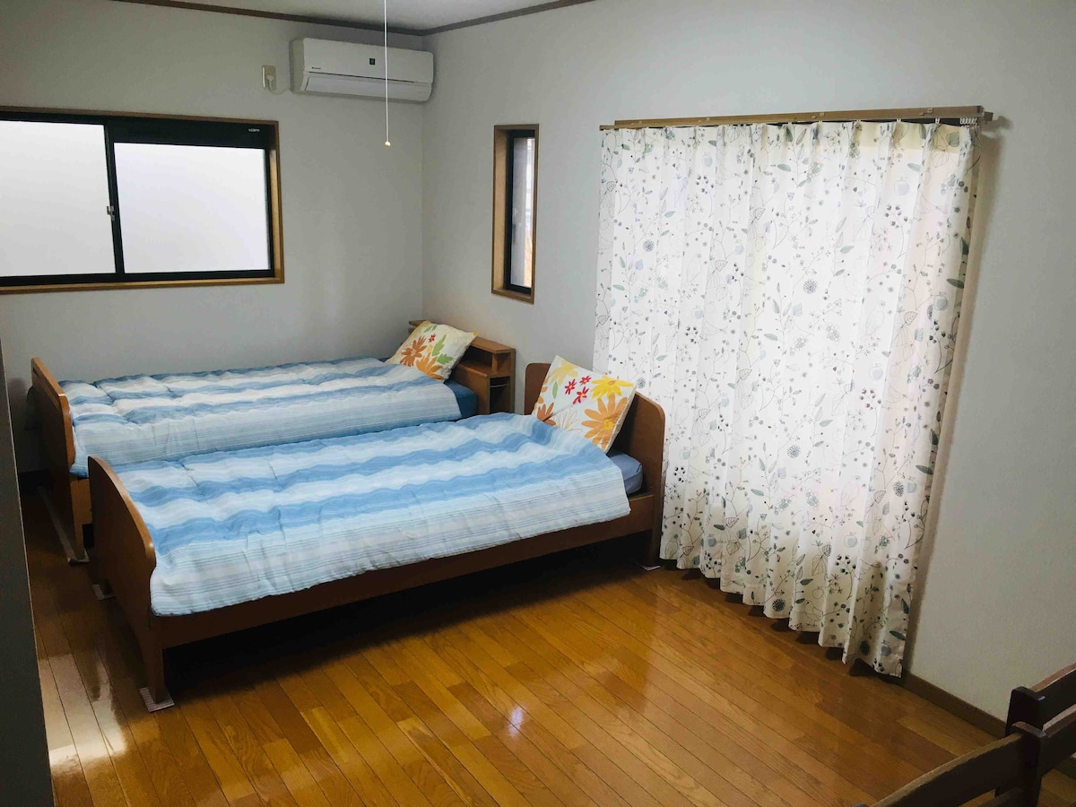 A well-lit room features two spacious beds positioned parallel to each other, with blue bedding and decorative pillows. A window adorned with sheer curtains allows natural light to filter in, while wooden floors add warmth to the space. A small bedside table stands between the beds.