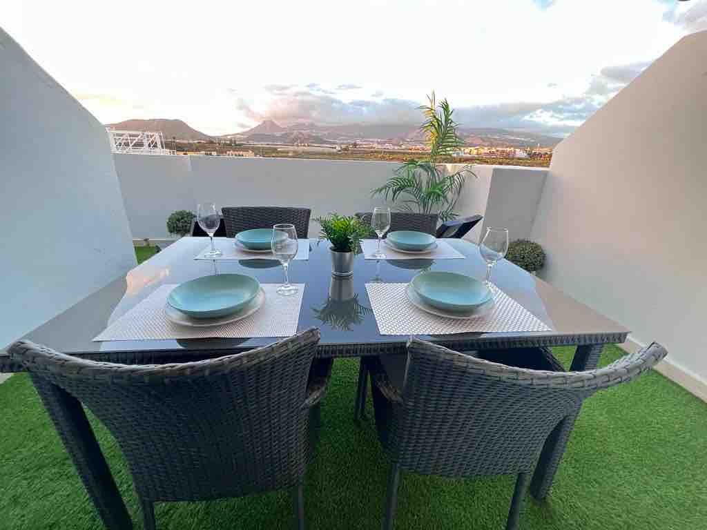 An outdoor dining area is presented with a glass table, surrounded by four wicker chairs. Each place setting includes a light blue plate and a wine glass. A small plant centerpiece adds a touch of greenery, with scenic views of the mountains in the background.