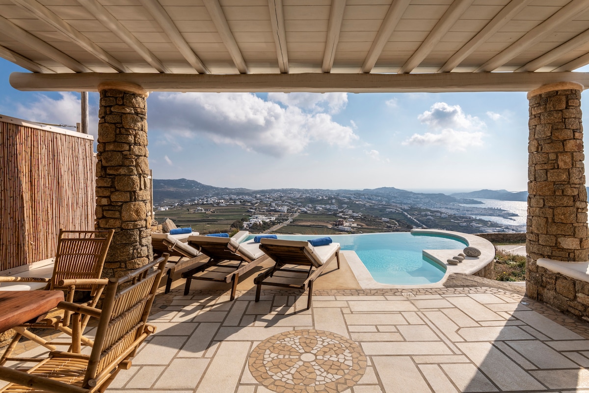 An outdoor terrace is framed by two stone columns, overlooking a private swimming pool and panoramic views of the Aegean Sea. Several wooden lounge chairs are arranged around a table on a patterned stone floor, creating an inviting space for relaxation.