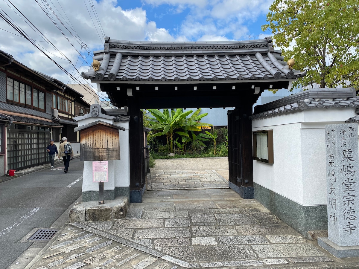 A traditional gateway is framed by stone pathways leading to a serene courtyard adorned with greenery. The structure features a tiled roof and wooden accents, while neighboring buildings reflect local architectural style. Clear skies punctuate the scene, contributing to a peaceful atmosphere.