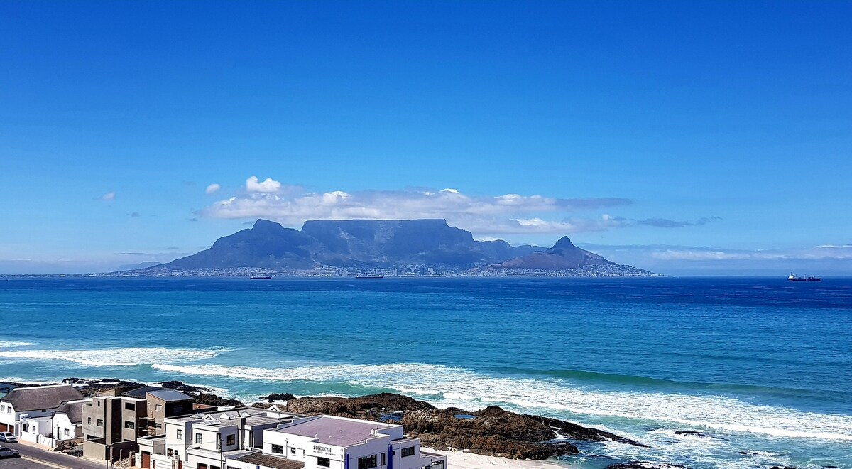 An expansive ocean view is captured, featuring the distinctive silhouette of Table Mountain against a clear blue sky. Gentle waves lap at the sandy shore, while a few boats can be seen on the horizon, completing this scenic portrayal of coastal life.