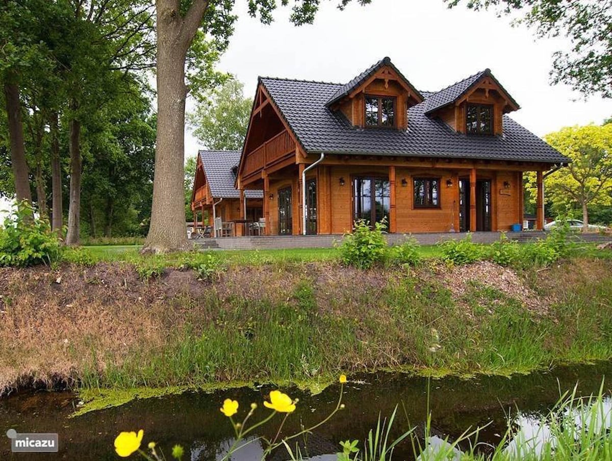 A wooden villa is surrounded by lush greenery, with a sloped roof and large windows. The exterior showcases a blend of natural wood tones and dark roofing. A tranquil pond is visible in the foreground, complemented by wildflowers and grass along the water's edge.