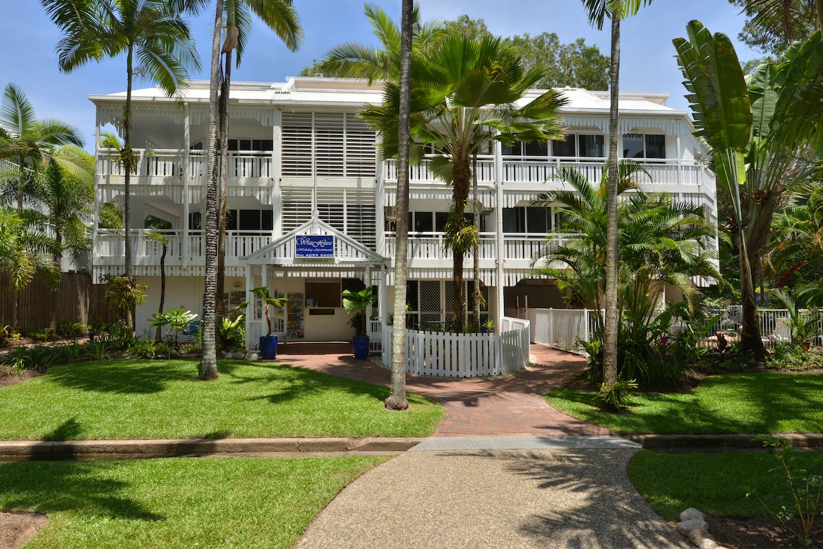 A Queenslander style building is surrounded by lush tropical landscaping, featuring a bright white façade and expansive wraparound verandas. Palm trees and vibrant foliage are present in the garden area, contributing to a serene and inviting setting near the park.