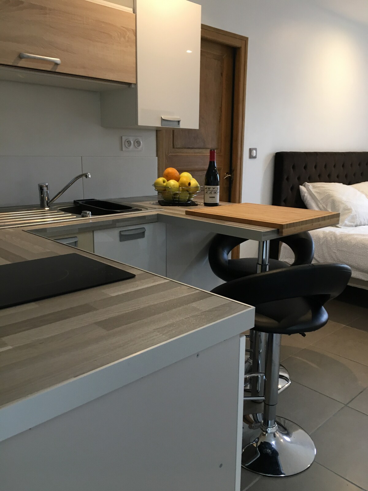 A modern kitchenette features a sleek countertop, a stainless steel sink, and a small dining area with high-backed stools. A bowl of fresh fruit and a bottle of wine are positioned on the countertop, with a glimpse of the spacious bed visible in the background.