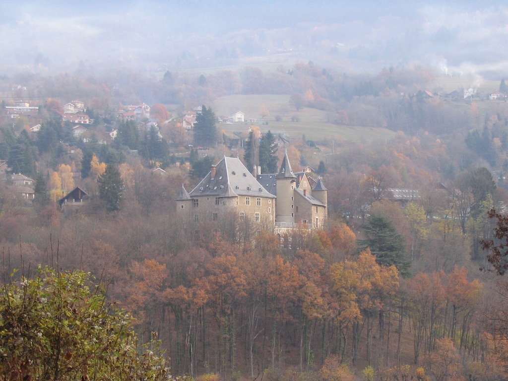 A historic castle is nestled among vibrant autumn foliage, with trees displaying hues of orange and gold. The castle's turrets and stone walls are gently enveloped by a misty landscape, set against rolling hills that extend into the distance under a soft sky.