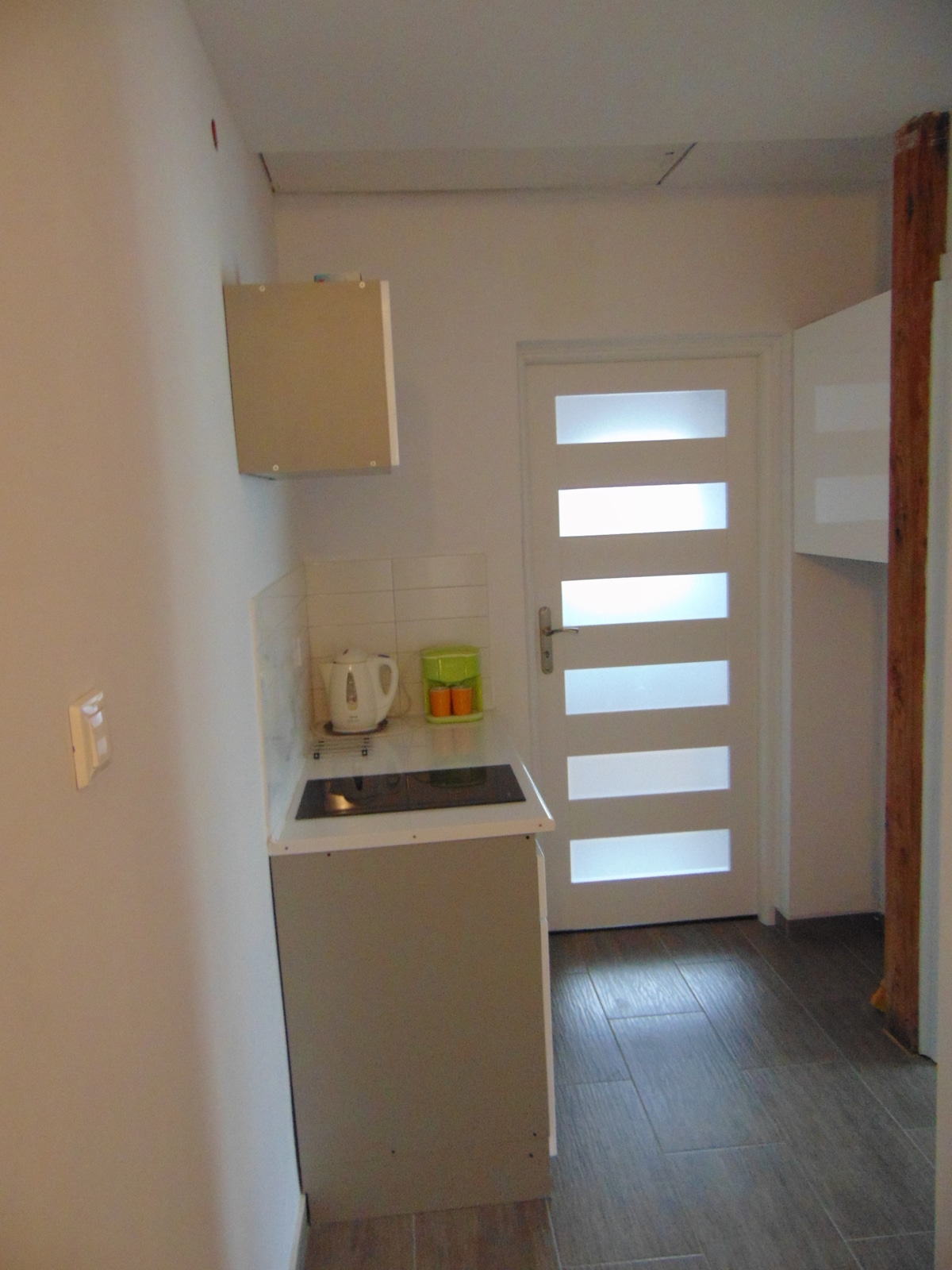A compact kitchen area features a modern electric stovetop and a kettle. Storage cabinets are positioned above the countertop, while a door with frosted glass panels allows natural light to enter. The floor is tiled with dark wood-like planks, enhancing the contemporary appearance.