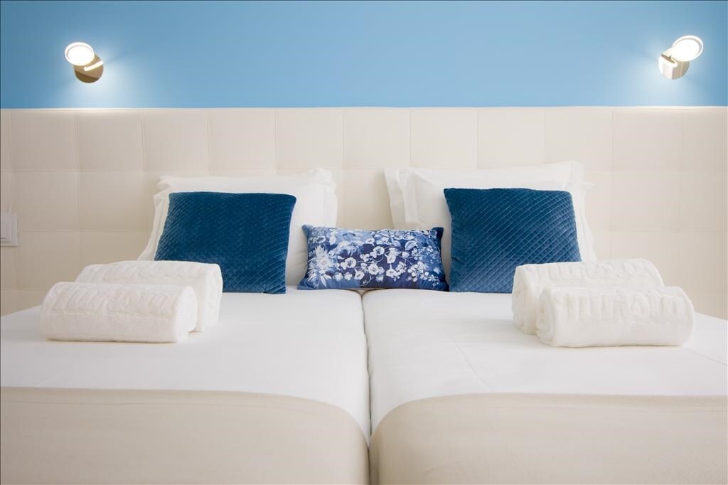 The image showcases two twin beds with a neutral-colored base, fitted with white linens and decorative pillows in shades of blue. Soft lighting is provided by wall-mounted fixtures on either side of the headboard, highlighting the calming blue wall behind.