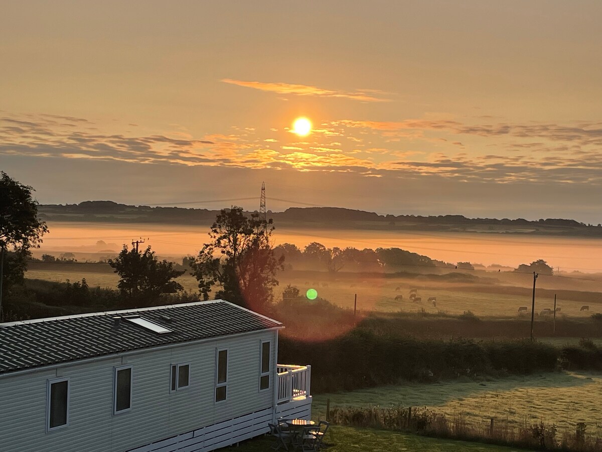 A tranquil scene captures the sun rising above a misty landscape. A mobile home is positioned in the foreground, showcasing a deck. Gentle rolling hills are visible in the distance, with soft clouds illuminated by the sun's warm glow.