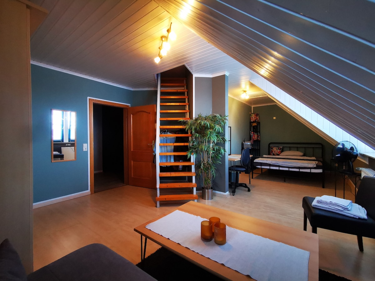 A spacious room is highlighted by wood flooring and a slanted ceiling. A comfortable seating area is visible, along with a dining table. A set of wooden stairs leads to an upper level, while a sleeping area with a bed is positioned in the corner. Natural light enters through multiple windows.