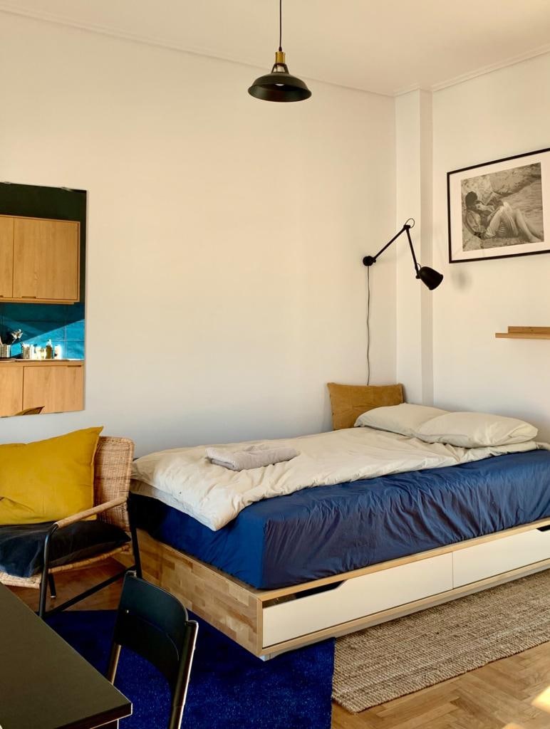 A compact and airy room features a spacious bed with a blue duvet and neatly arranged towels. A cozy armchair and a small dining table add functionality. Natural light filters through the window, highlighting the minimalist decor, including shelving and framed artwork.