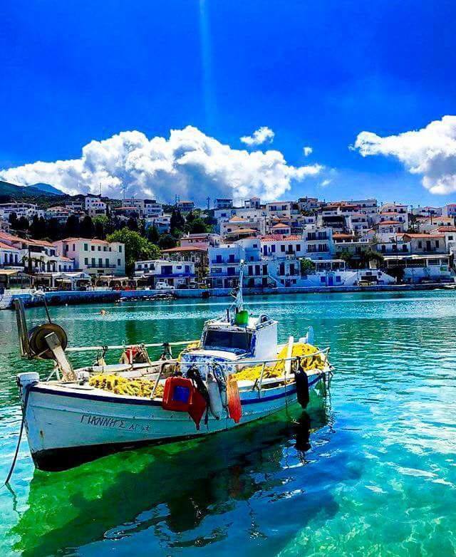 A traditional fishing boat is anchored in calm, clear waters, surrounded by brightly colored buildings of a seaside village. The blue sky features scattered clouds, reflecting the vibrant atmosphere of the area.