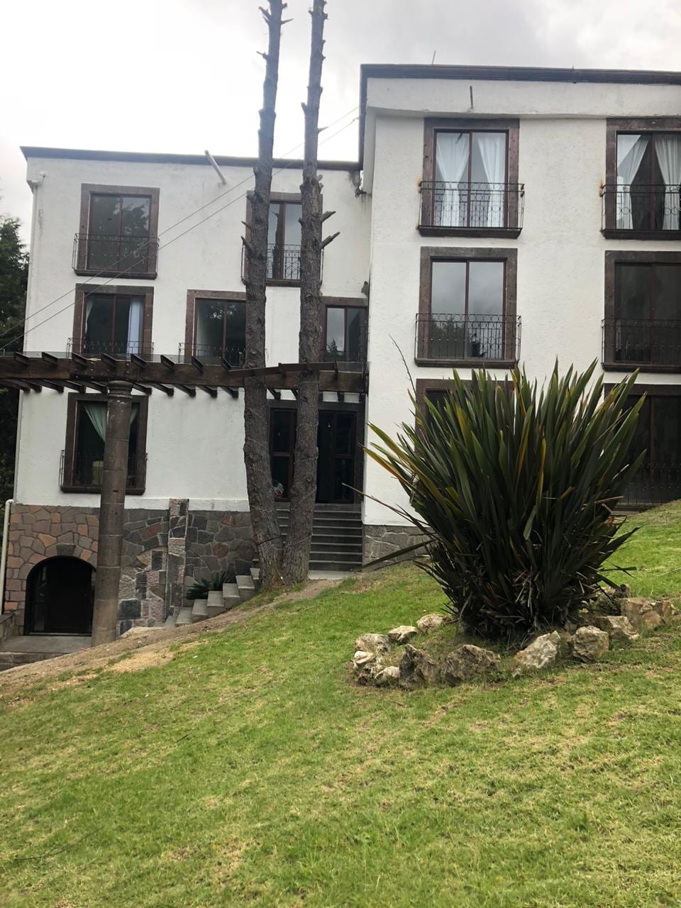 The exterior of a multi-story building is visible, featuring a mix of white walls and stone accents. Several large windows are adorned with black railings, and a stairway leads to the entrance. Lush greenery surrounds the structure, enhancing the natural setting.